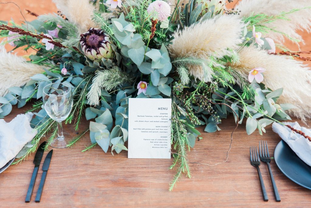 Bohemian pampas and protea centrepiece and menu on a picnic style wedding table styled by Oh Happy Day in South Africa