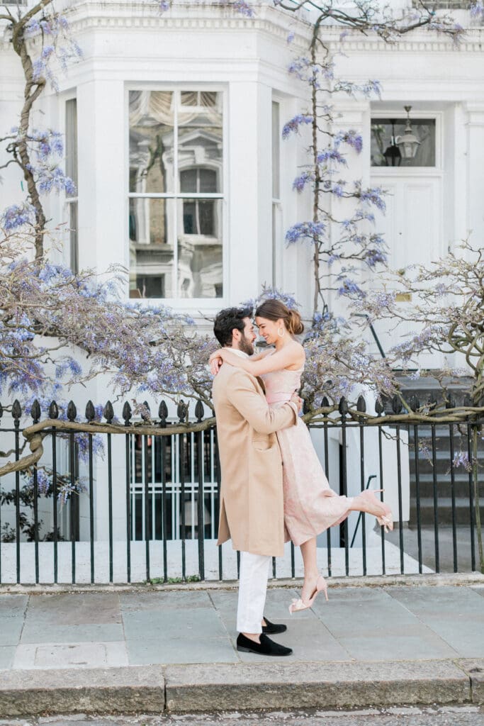 A romantic couple posting for their engagement shoot with a backdrop of wisteria behind them for their engagement shoot in London. The groom is lifting his bride off the floor in a cute pose.