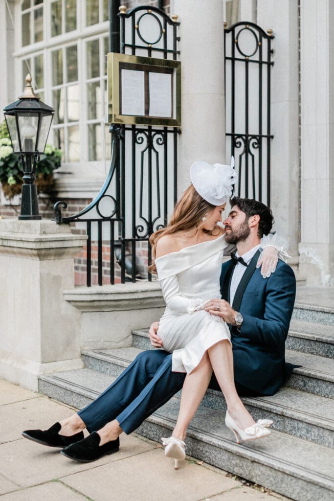 Bride sitting on grooms lap in an romantic embrace whilst the groom is sitting on the outside steps of The Belgravia, London, following their wedding ceremony.