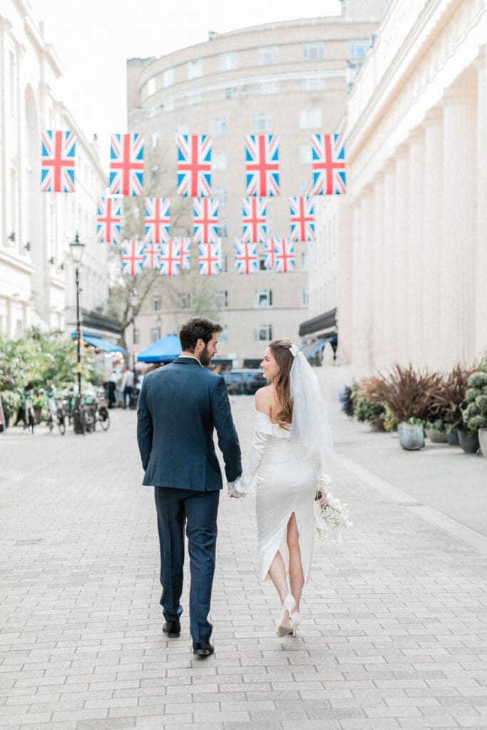 Bride and Groom walking along the streets of London, away from the camera with Union Jack flags above them.