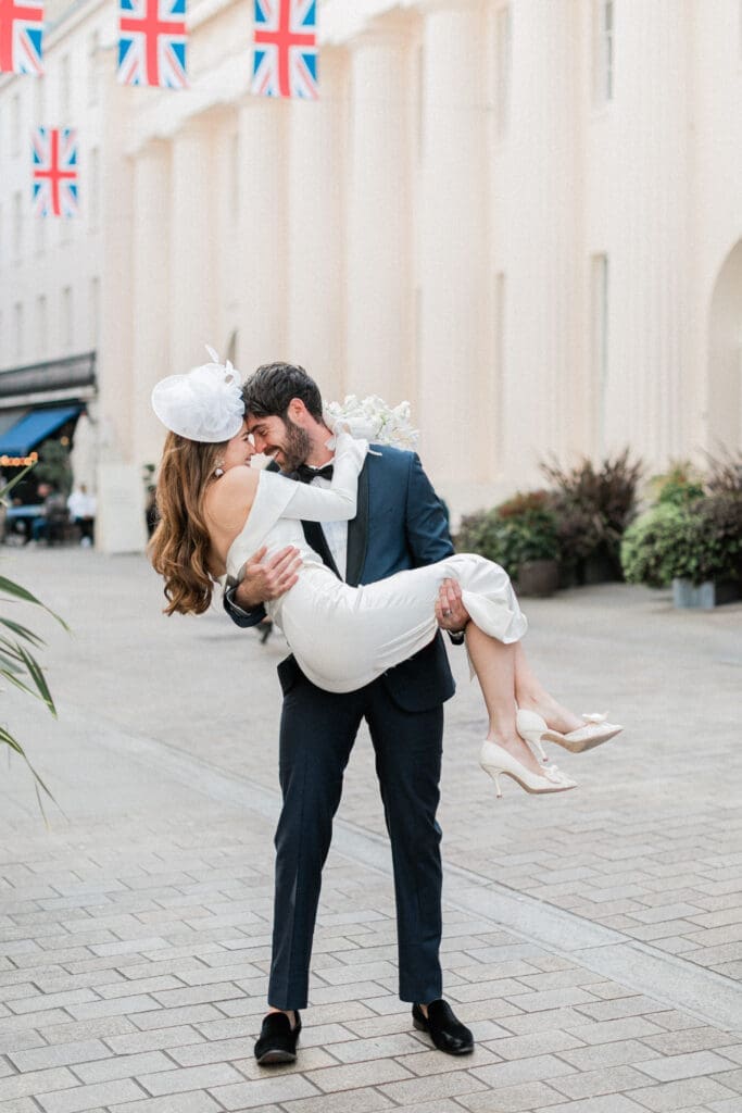 A groom picking up and holding his bride on the streets of Chelsea in London with Union Jack flags above them.