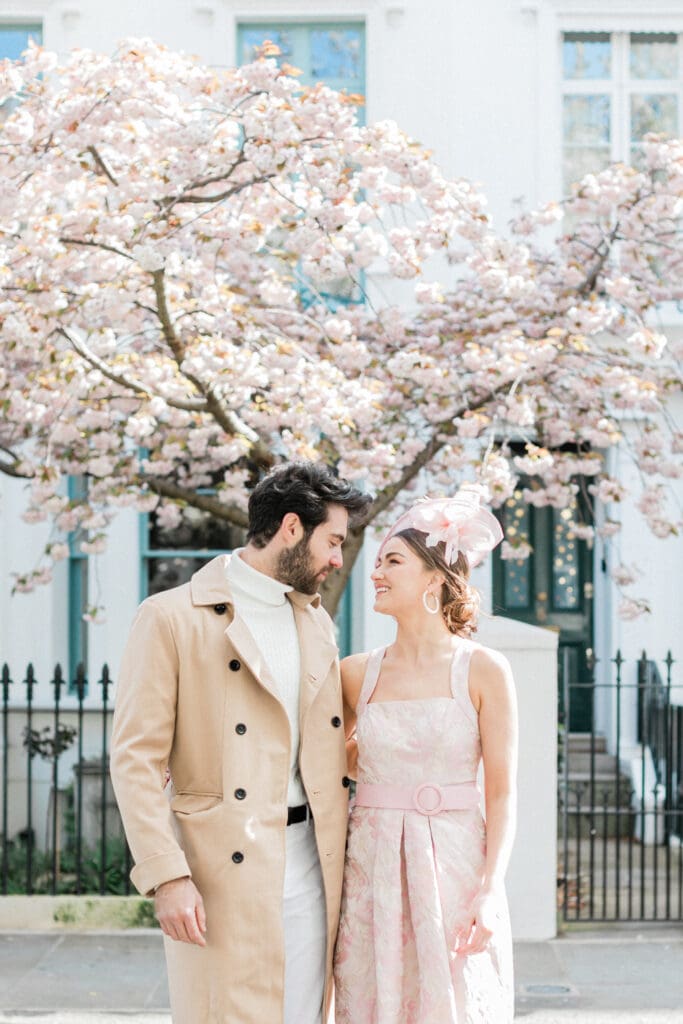 A couple posing outside a white townhouse for their engagement shoot in Notting Hill with a pink blossom tree behind them.