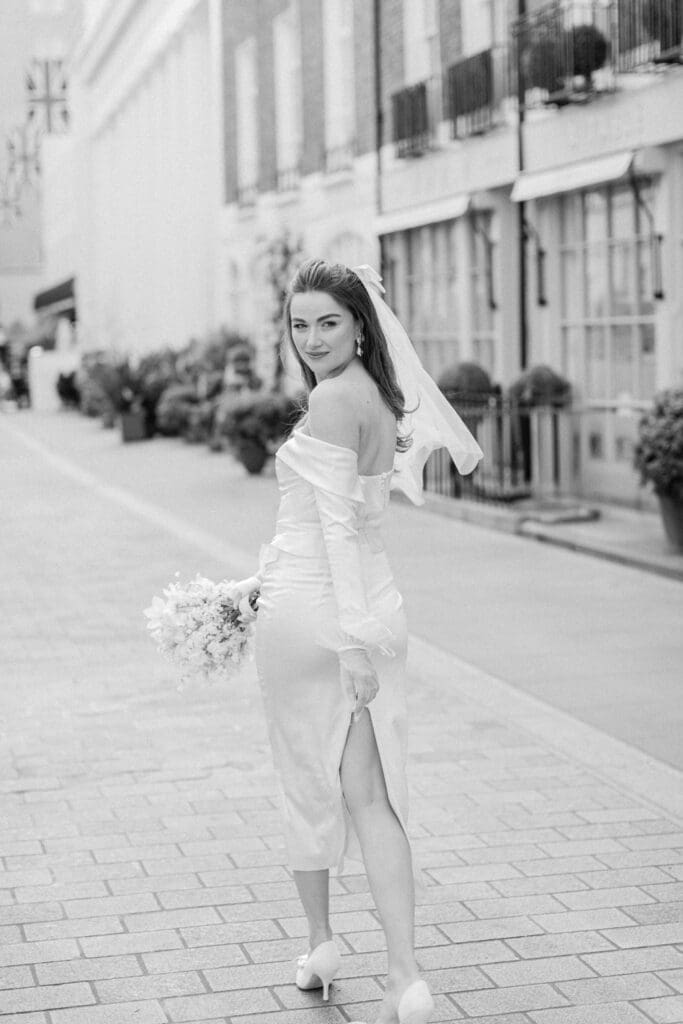 Black and white wedding photo of the bride walking along a street in Chelsea whilst looking back at the camera with her bridal bouquet in hand.