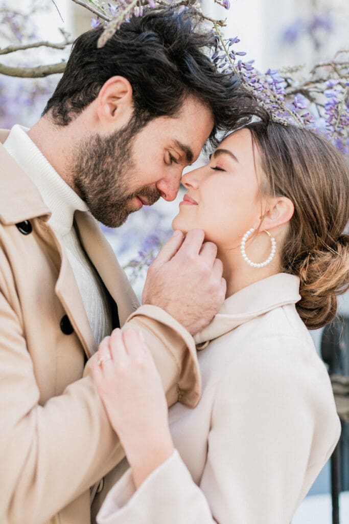 A romantic couple leaning into each other face to face with purple wisteria behind them in Notting Hill