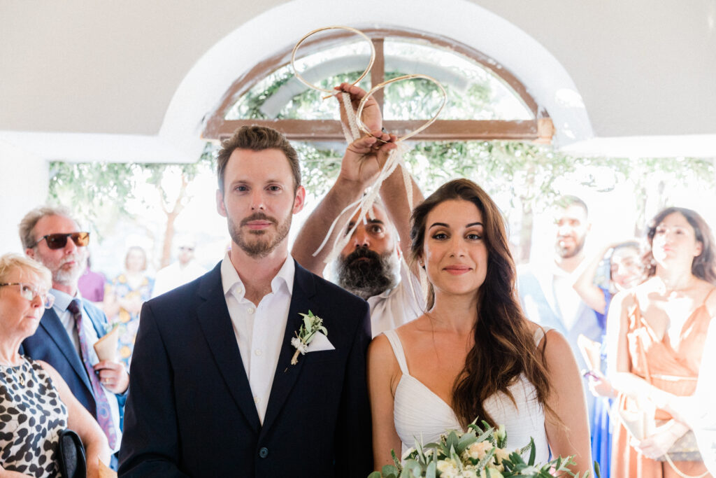 The bride and groom looking straight into the camera with the priest behind them performing traditional greek wedding practices.