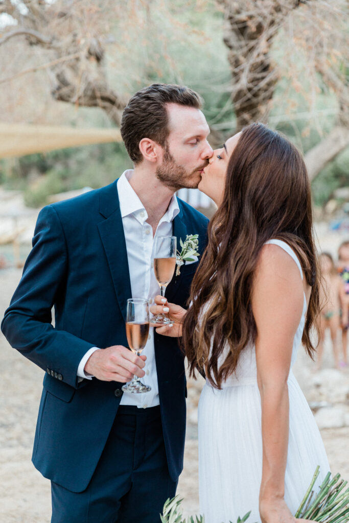 Bride and Groom sharing a kiss whilst celebrating their wedding reception and holding a glass of champagne each in Ithaca, Greece.
