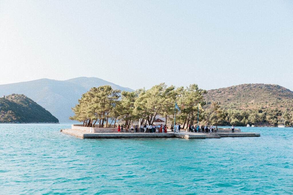 The island in Ithaca, greece, where the ceremony takes place. It is accessible only by boat. The guests are standing on the island, waiting for the bride and groom to arrive for the ceremony.