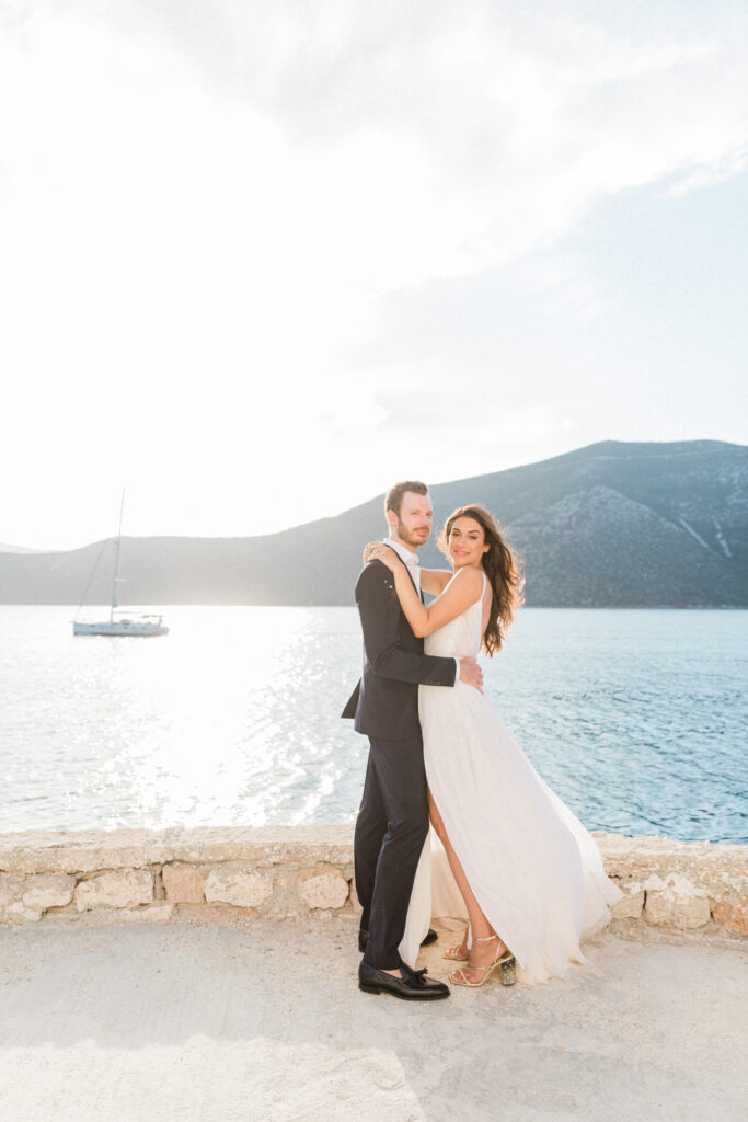 The bride and groom embracing each other on the beach whilst looking at the camera with the sea and a boat in the background at their wedding in Greece.